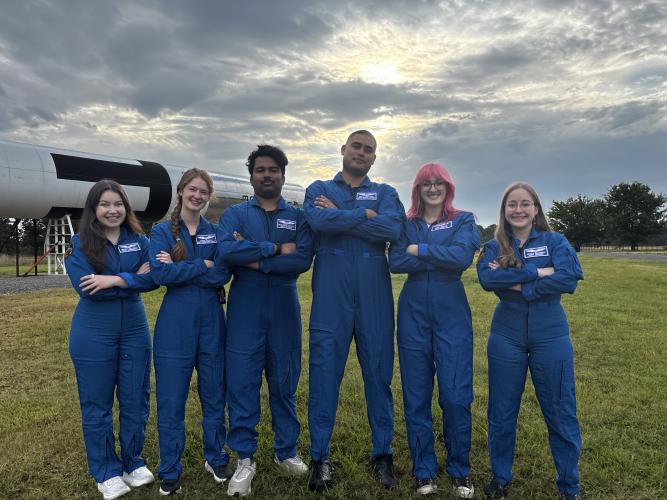 Five people wearing matching blue flight suits stand with arms crossed in a grassy field beside a large rocket display. Sunlight breaks through clouds overhead in the background.